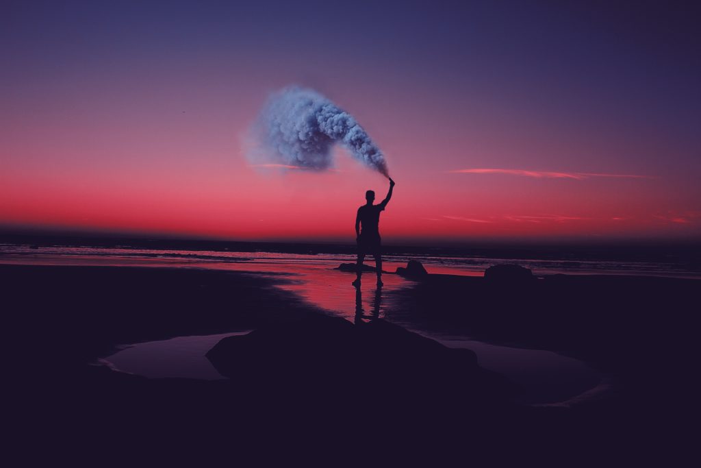 silhouette of man standing on seashore holding smoke can